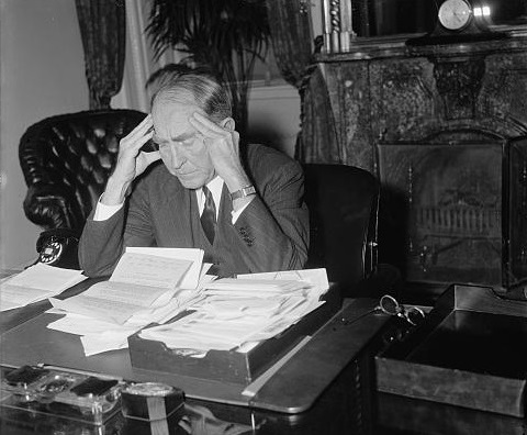 Black-and-white historical photograph of a man seated at a desk covered with papers, holding his hands to his temples as if overwhelmed.