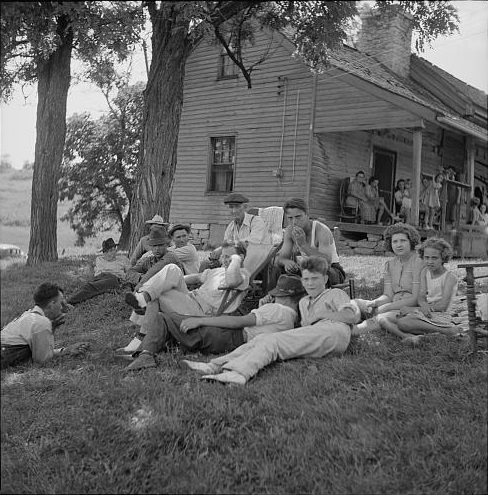 Black-and-white historical photograph of a large group of people gathered outside a farmhouse, with several people sitting in the grass beneath a tree and others standing or sitting on the porch.