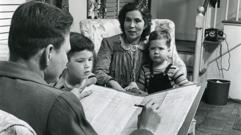 Black-and-white historical photograph of a census enumerator interviewing a mother and her two young sons, with a large census form visible in the foreground.