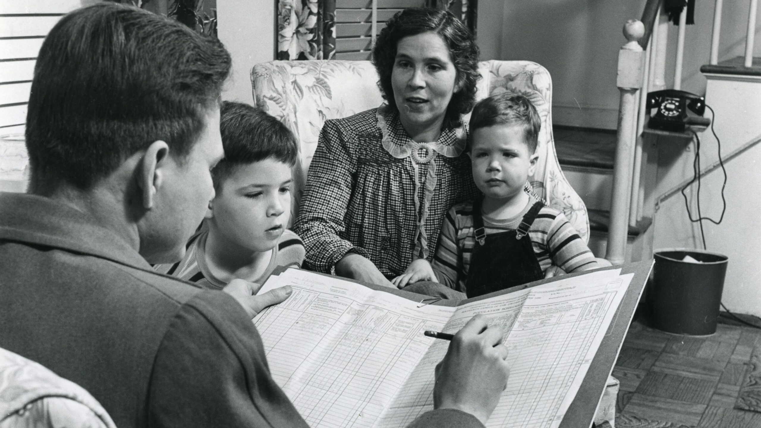 Black-and-white historical photograph of a census enumerator interviewing a mother and her two young sons, with a large census form visible in the foreground.